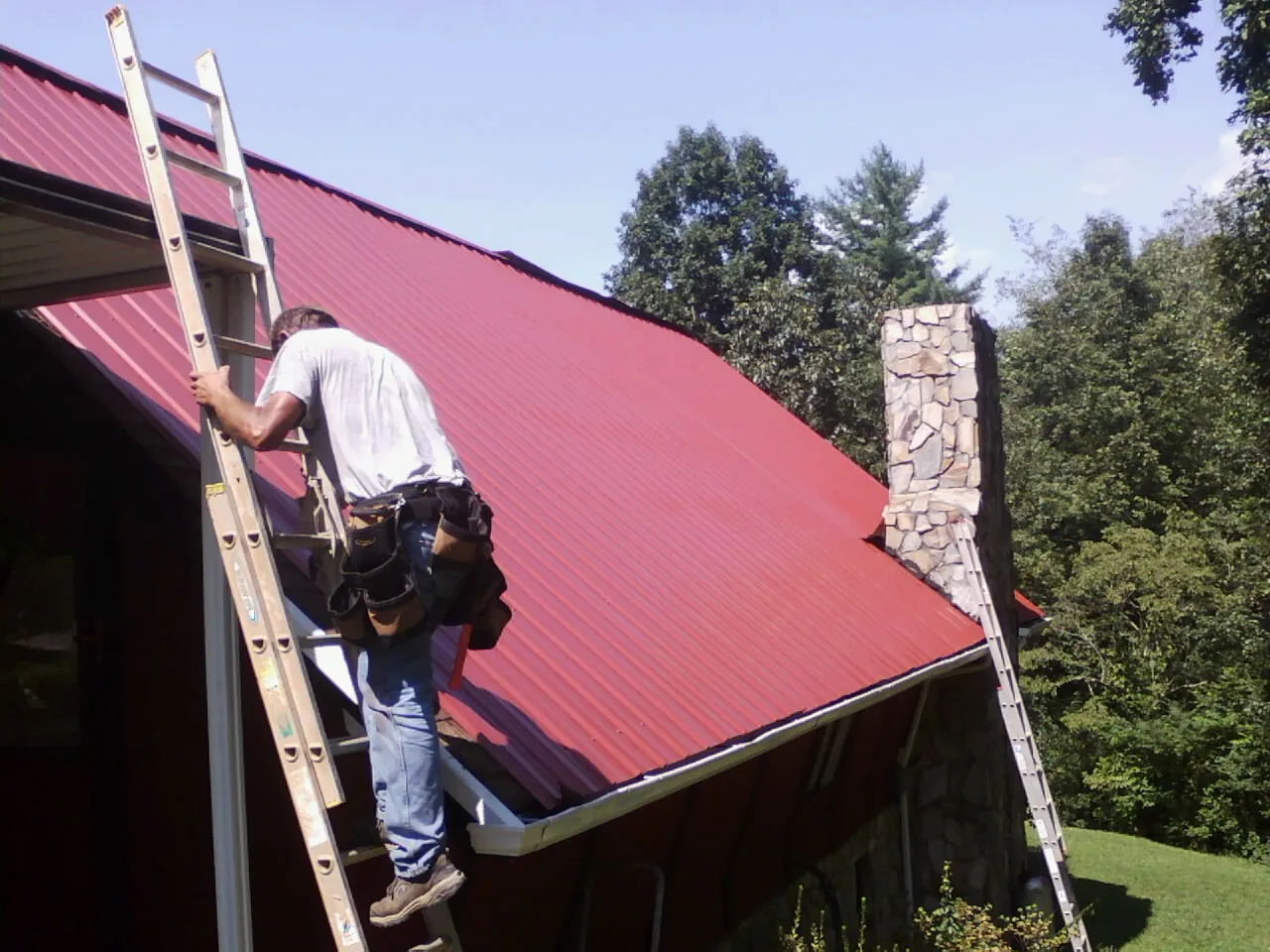 Roofer on a ladder beside a red metal roof and stone chimney — Soffit & Fascia Repair by Roofing Brothers in Mount Vernon